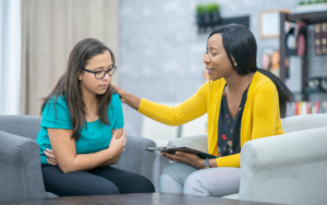 On the right, a counselor in a yellow sweater comforts a young woman on the left sitting on armchairs..