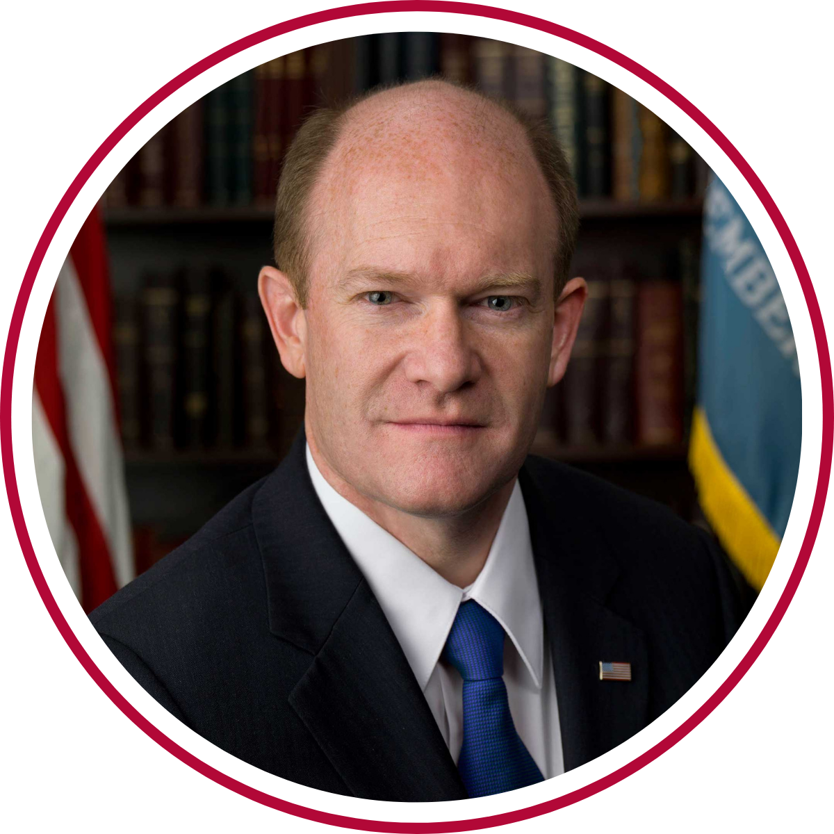 Professional headshot of a man wearing a dark suit, white shirt, and blue tie with a U.S. flag pin on the lapel. Bookshelves and partially visible flags appear in the background. The portrait is framed within a circular border.