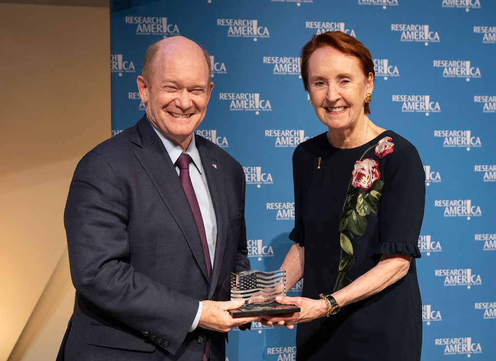 A man and a woman--Senator Chris Coons and Mary Woolley—stand on a stage in front of a blue backdrop repeatedly printed with the “Research!America” logo. They are smiling while jointly holding a clear glass award shaped like a waving American flag. The man wears a dark suit with a white shirt and burgundy tie, and the woman wears a dark dress with a floral design on the shoulder. The photo appears to capture the moment an award is being presented.