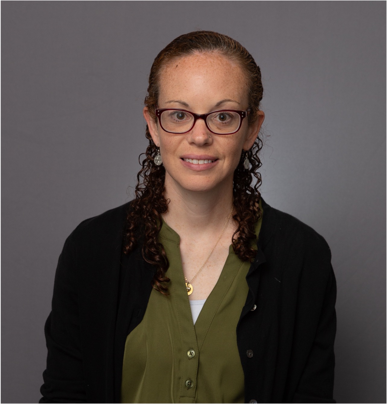 Woman with curly hair and glasses, wearing a green blouse and black cardigan, smiles slightly against a plain gray background. Professional tone.
