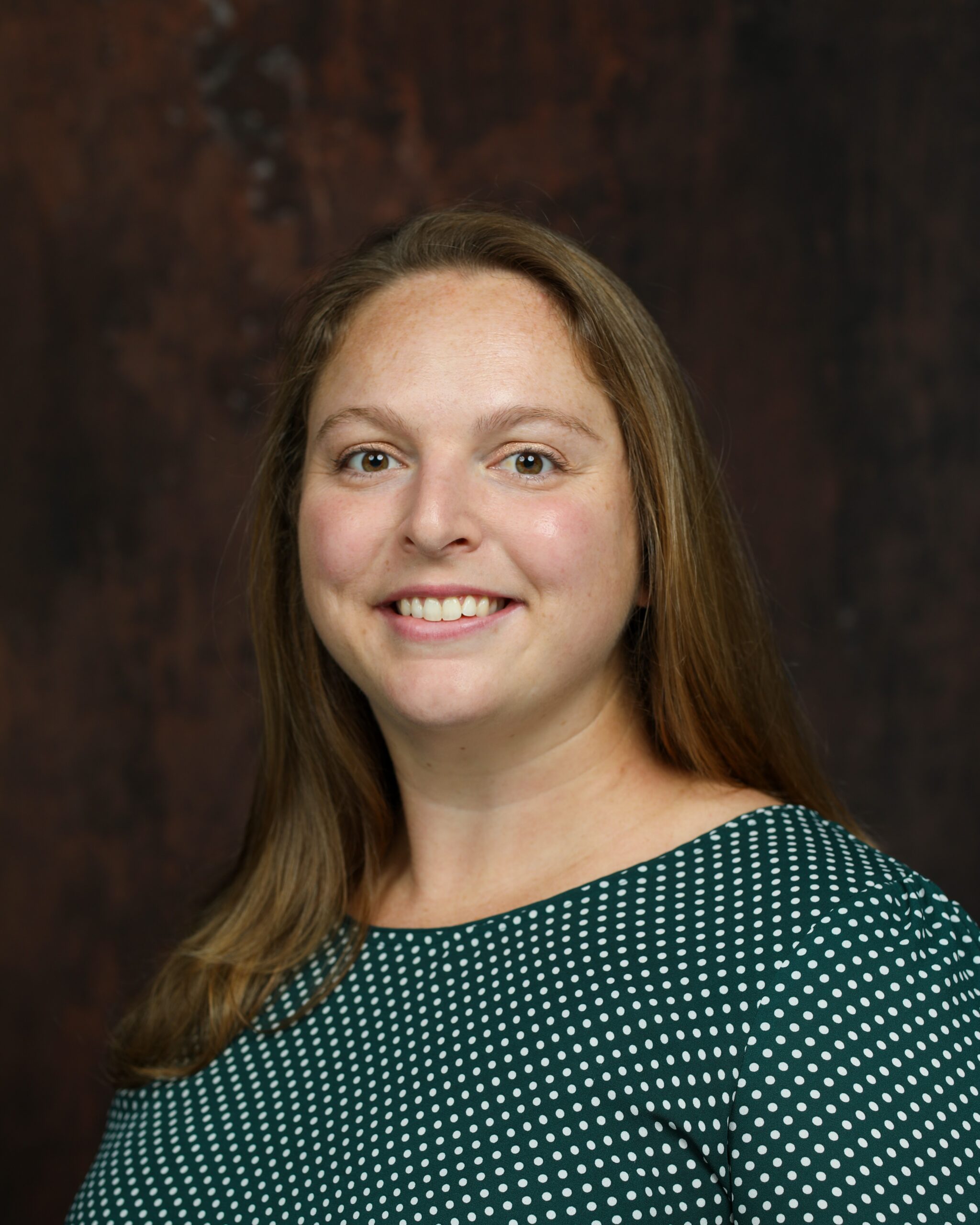 A woman with long brown hair smiles warmly against a dark, textured background. She wears a green polka dot top, conveying a friendly and approachable tone.