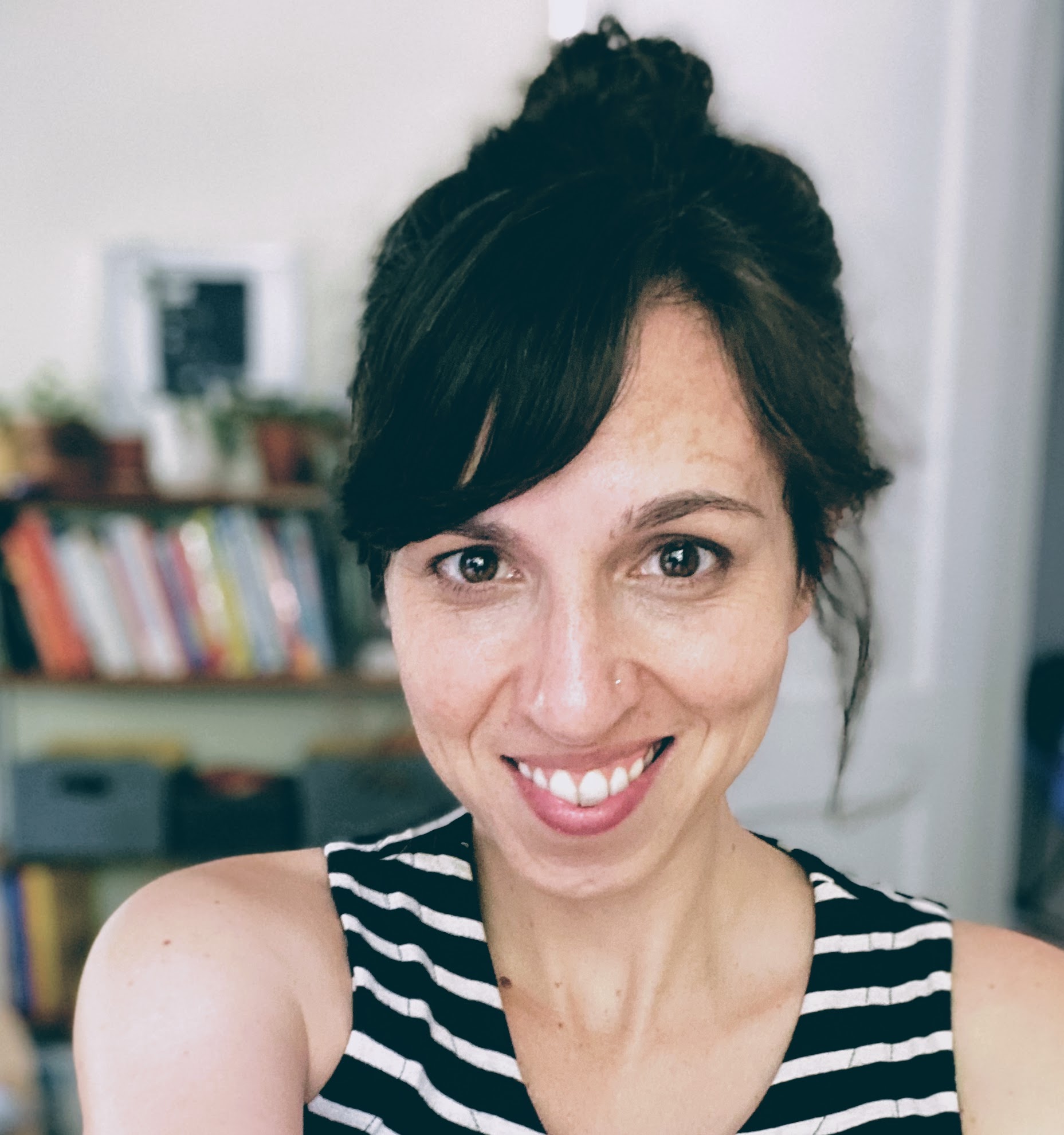 Smiling woman with dark hair in a bun, wearing a striped top. Background shows blurred books and plants, conveying a cozy, cheerful atmosphere.